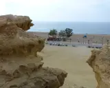 Rocas erosionadas en primer plano, con la playa de Bolnuevo, el mar y un paseo marítimo con palmeras al fondo.