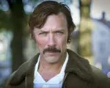 Porträtt av en svensk manlig skådespelare från 2000-talet med mustasch och blå ögon, klädd i en brun jacka med fuskpäls.
