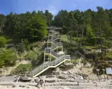 Długa, drewniana klatka schodowa prowadzi na szczyt kawczej góry. Na plaży siedzi para z psem.