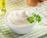 Un bol de mayonnaise maison onctueuse, garni de persil frais. Des œufs et un fouet à proximité suggèrent la préparation.