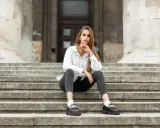 Eleganckie mokasyny to idealne buty do szarych spodni. Kobieta w białej koszuli i czarnych jeansach siedzi na schodach.