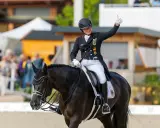 Reiterin gibt Daumen hoch nach erfolgreichem Grand Prix Reiten. Dunkles Pferd mit Turnieroutfit und Helm.