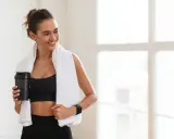 Lächelnde Frau mit Handtuch und Shaker nach dem Training. Sie fragt sich, wie viel Kreatin pro Tag für optimale Ergebnisse.
