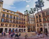 Sonniger Platz in der **Malaga Altstadt** mit einem Brunnen, Palmen und bunten Gebäuden. Menschen flanieren und genießen die Atmosphäre.