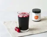 Ein Glas dunkler Sauerkirschsaft mit Eiswürfeln und zwei Kirschen daneben. Eine Dose mit der Aufschrift "Ganz Gelassen" steht im Hintergrund. Die positive sauerkirschsaft wirkung wird hier visuell dargestellt.