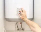 Mano ajustando el control de temperatura de un termo eléctrico. La instalación se ve limpia y moderna.