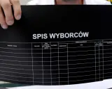 Ręce trzymają czarny dokument z napisem "SPIS WYBORCÓW". Widoczne kolumny do uzupełnienia, sugerujące dopisanie do spisu wyborców.