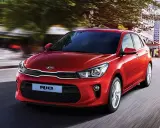 Un hatchback rojo Kia Rio se desplaza por una calle urbana, con edificios y árboles borrosos por el movimiento.