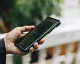 Dłoń trzyma mokry smartfon ip67 na tle zieleni. Krople deszczu spływają po ekranie.