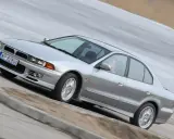 Srebrny Mitsubishi Galant z gracją pokonuje zakręt. Kierowca w okularach pewnie prowadzi auto.