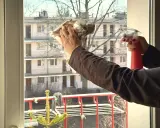 Ręce czyszczą okno, jedna trzyma spryskiwacz, druga zgniata gazetę do polerowania. Skuteczne mycie okien octem to sekret czystości.