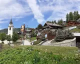 A picturesque village in Chamois, Italy, with a church tower, wooden chalets, and lush greenery. Scopri cosa vedere in questo incantevole borgo alpino.