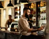 Hombre con barba gestionando un bar, usando una tablet y una pantalla táctil para procesar pagos.
