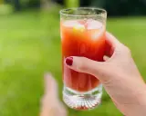 A hand with red nail polish holds a tall glass of Bloody Mary with ice and a cucumber slice, outdoors.