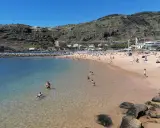 Sonniger Strand in Machico, Madeira, mit vielen Menschen, die im klaren Wasser planschen und sich am Sandstrand sonnen.