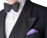 A man wears a black tuxedo with a pleated white shirt and a black bow tie. A purple pocket square adds a pop of color. This outfit answers the question: can you wear a tie with a tux? (No, a bow tie is traditional.)