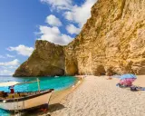 Ein Boot liegt am Korfu Strand vor einer beeindruckenden Felswand. Türkises Wasser und ein sonniger Himmel laden zum Verweilen ein.