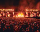 Un'immensa folla partecipa a un festival tekno, illuminata da fiammate spettacolari e schermi con immagini di fuoco.