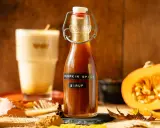 Flasche mit goldbraunem Pumpkin Spice Sirup, daneben ein Latte Macchiato, Kürbishälften und Herbstlaub.