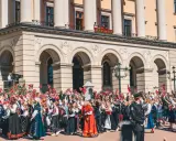Tłum ludzi, w tym dzieci, świętuje, machając norweskimi flagami. Wiele osób nosi tradycyjny strój norweski, ozdobiony haftami i kolorowymi wzorami.