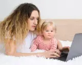 Kobieta z dzieckiem na rękach pracuje na laptopie, zastanawiając się, czy na urlopie wychowawczym można pracować.