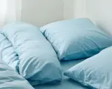 En bädd med ljusblå sängkläder, troligen percale eller satin, med flera kuddar och ett täcke.