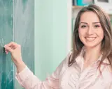 Uśmiechnięta nauczycielka pisze kredą na tablicy. Marzy o tym, by podwyżki dla nauczycieli 2026 przyniosły jej zasłużone docenienie.