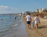 Rodzina spaceruje po piaszczystej plaży w Durrës. W tle widać morze, budynki i parasole plażowe.