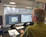 Mann am Computer, der Baupläne nach DIN 4426 erstellt. Auf dem Bildschirm sind CAD-Zeichnungen und eine Liste von Bauteilen zu sehen.