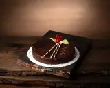 En lyxig fransk chokladtårta med chokladbitar, en röd ros och vita chokladpinnar, serverad på en träplatta.