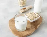 Ein Glas cremiger Hafermilch steht auf einem Holzbrett, daneben Schüsseln mit Haferflocken. Hafermilch gesund und lecker!