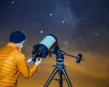 Mężczyzna w pomarańczowej kurtce ustawia aparat Canon EOS Ra do astrofotografii pod rozgwieżdżonym niebem.
