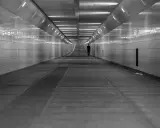 Człowiek w garniturze idzie przez długi, pusty tunel. Światła na ścianach tworzą wrażenie nieskończoności, jakby wchodził w szarą strefę.