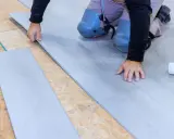 Man con rodilleras coloca cuidadosamente tablones de suelo laminado gris sobre una base de madera.