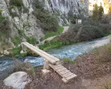 Puente de madera sobre un río caudaloso, parte de la ruta del zarzalar. Senderos y vegetación agreste flanquean el camino.