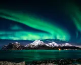 Zielona zorza polarna nad ośnieżonymi górami i morzem. Marzysz, jak dojechać do Norwegii, by zobaczyć to na żywo?