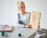 Kobieta prezentuje obraz z nici i gwoździ szablon. Na stole widać inne prace i materiały do tworzenia.