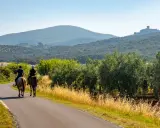 Due cavalieri godono di una **passeggiata a cavallo toscana** tra ulivi e colline, con un castello in lontananza.