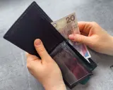 Ręce wkładają banknot 20 zł do portfela. Dowiedz się, komu przysługuje zasiłek dla bezrobotnych.