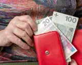 Dłoń starszej osoby wkłada pieniądze do czerwonego portfela. Może to być dodatek mieszkaniowy.