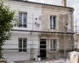 Deux ouvriers sur des échafaudages sont en train de refaire sa façade, appliquant une nouvelle couche de peinture blanche sur un mur décrépi.