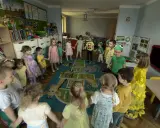 Dzieci trzymają się za ręce, tworząc krąg na dywanie z zabawnymi zwierzątkami. To radosna zabawa w **zespole szkolno przedszkolnym**.