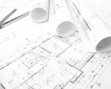 Projekt budowlany zawiera plany pomieszczeń, wymiary i detale, jak okna. Cyfrowy cyrkiel leży obok zwiniętych rysunków.