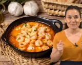 Una mujer sonríe mientras muestra cómo hacer gambas al ajillo, con un plato humeante y los ingredientes listos.