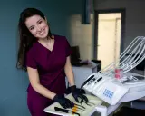 Uśmiechnięta asystentka stomatologiczna w bordowym uniformie przygotowuje narzędzia na tacce obok fotela dentystycznego.