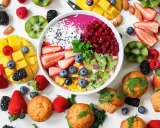 Kolorowa miska smoothie z owocami, jagodami, granatem i wiórkami kokosowymi. Obok mango, truskawki, kiwi, maliny i muffinki. Idealne owoce na noc.