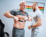 Mann legt Brustgurt für Muskelerhalt Training an, Trainer hilft.