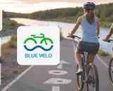 Para rowerzystów na ścieżce nad rzeką, obok logo "BLUE VELO".