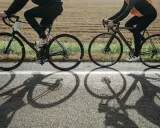 Dwie osoby na rowerach, w kaskach, jadące po drodze. Długie cienie rowerów na asfalcie.