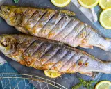 Zwei gegrillte Saiblinge liegen auf einem Steinplatte, garniert mit Zitronenscheiben und Kräutern. Perfekt für den Grill – wie lange dauert das?